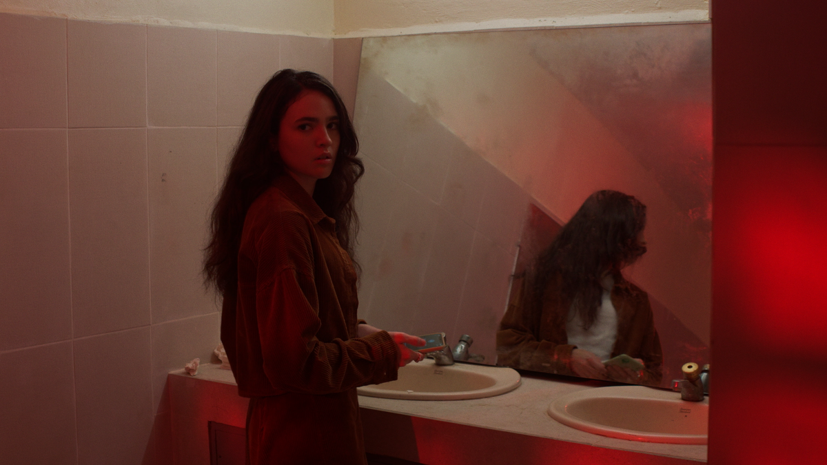 Liz, our protagonist, looks anxiously over her shoulder. She is in a dilapidated public bathroom and her reflection is visble in the dirty mirror.