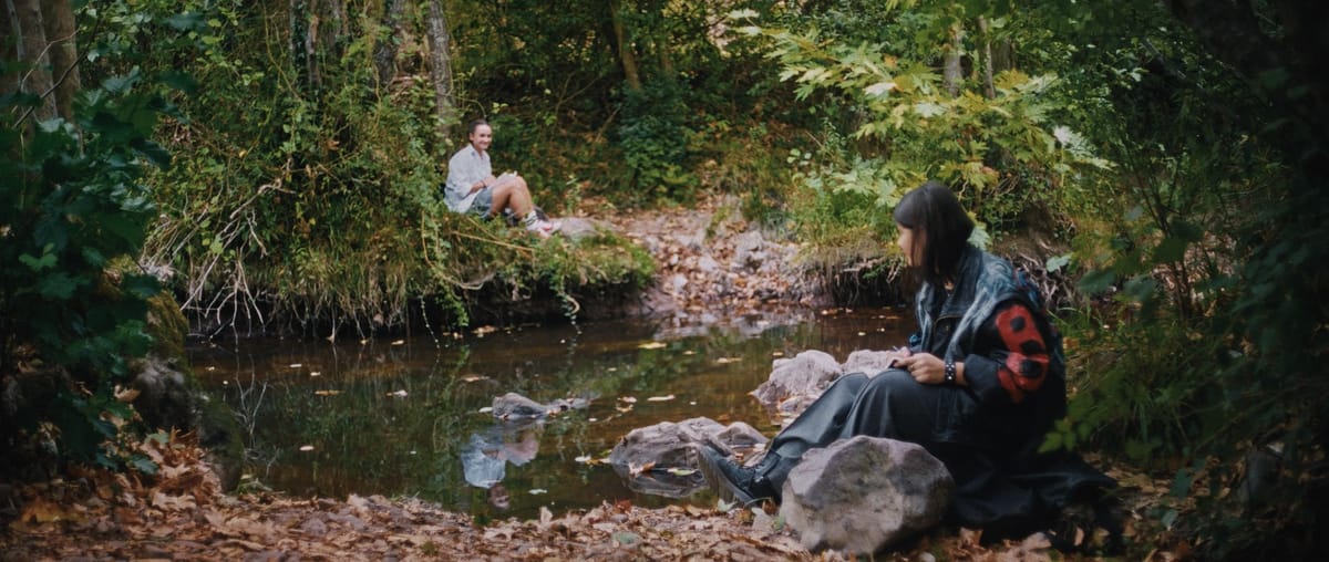 Two teenage girls, Ceren and Azra, sit across from each other at a verdant pond.