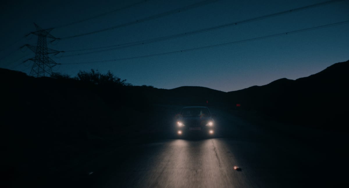 A car travels down a road at twilight with its headlights on, darkness surrounding it.