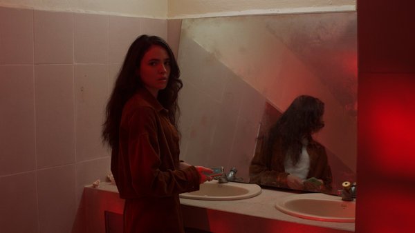 Liz, our protagonist, looks anxiously over her shoulder. She is in a dilapidated public bathroom and her reflection is visble in the dirty mirror.