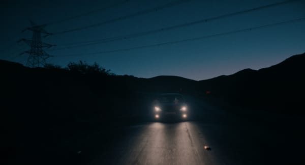 A car travels down a road at twilight with its headlights on, darkness surrounding it.